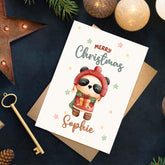 Christmas card with a panda character and 'Merry Christmas Sophie' text, surrounded by festive decorations on a dark surface.